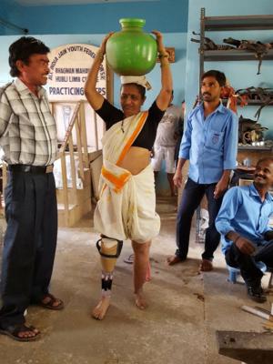 Indian woman with prosthetic leg walking with jar on her head while men watch